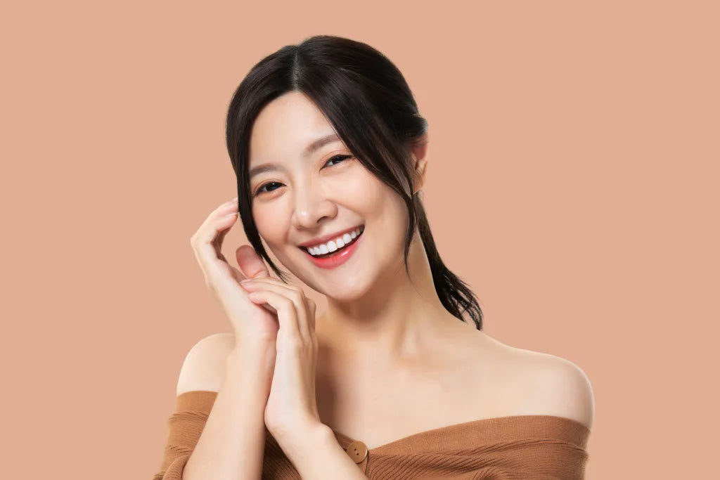 A smiling woman with dark hair wearing an off-the-shoulder brown top against a solid tan background.